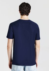 Uomo che indossa una semplice maglietta a maniche corte blu navy con risvolti e jeans in denim blu chiaro, visto da dietro.