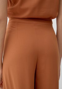 Pantalon en tissu lisse de couleur rouille avec une taille ajustée et des détails de coutures subtiles. Design fluide avec une coupe décontractée.