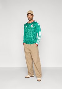 Man i grön Mexiko fotbolls-tröja, beige keps, lösa beige byxor och cremefärgade sneakers, stående med händerna i fickorna mot enfärgad bakgrund.