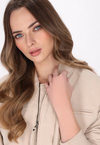 Giacca bomber beige con chiusura a zip, colletto a costine e tessuto testurizzato. I capelli ondulati del modello incorniciano delicatamente il viso, evidenziando occhi blu.