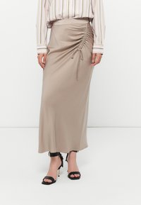 Beige Maxikleid mit seitlicher Raffung und Kordelzugsdetail, aus glattem Stoff gefertigt. Kombiniert mit schwarzen Riemchenabsätzen mit Knöchelriemen.
