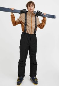 Tenue de ski comprenant une veste polaire zippée marron et un pantalon de neige noir avec des bretelles ajustables. Bottes de ski noires avec des accents jaunes.
