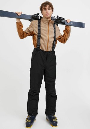 Skiing outfit featuring a brown zip-up fleece jacket and black snow pants with adjustable straps. Black ski boots with yellow accents.