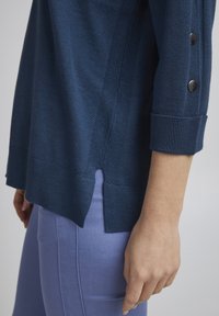 Marinefarbener Strickpullover mit gerippten Bündchen und Seitenschlitzen, versehen mit silberfarbenen Knopfakzenten an den Ärmeln. Kombiniert mit hell-lila Hosen.
