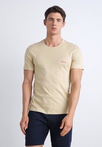 Beige bomull T-skjorte med korte ermer, rund hals, liten rød "HUGO"-logo på brystet, paret med marineblå shorts. Glatt tekstur.