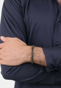 Braccialetto a catena nera su un polso, con anelli interconnessi e accenti arrotondati, abbinato a una camicia scura con colletto.