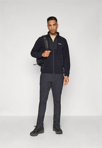 Czarna kurtka z polaru na zamek, w zestawie z czarnymi spodniami i butami trekkingowymi. Modelka niesie plecak i stoi na neutralnym tle.