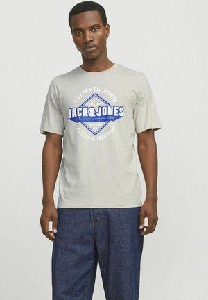 Svijetlo siva pamučna majica s kružnim plavim i bijelim grafičkim logotipom koji glasi "AUTENTIČNI DENIM JACK & JONES", uparen sa tamnim trapericama.