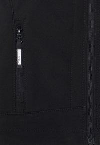 Veste softshell noire avec une poche zippée, texture lisse et détails de couture le long des coutures. Logo visible sur le tirette du zip.