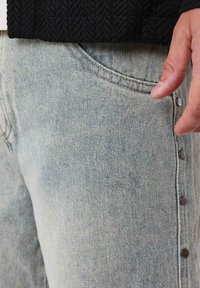 Vaqueros de mezclilla azul claro con un patrón texturizado, que presentan un cierre lateral con botón y un bolsillo frontal. El tejido parece suave y desgastado.