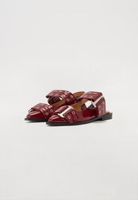 Sandales plates en cuir verni bourgogne avec des bouts pointus, larges sangles avec des œillets en argent et des boucles sur un fond blanc.