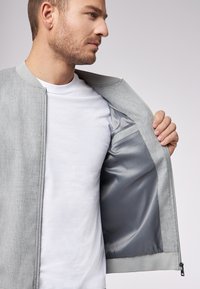 Giacca bomber grigia chiara con una texture liscia e polsini a costine, aperta per rivelare una fodera interna grigia simile alla seta. T-shirt bianca sotto.