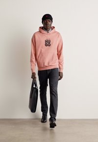 Sudadera rosa claro con la impresión negra "HUGO", pantalones gris oscuro y zapatos negros, complementados con una bolsa de cuero negra y un gorro.