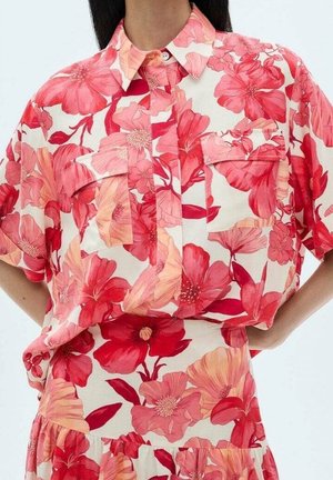 Vrouw die een rood, roze en perzikkleurig bloemig kortemouwenhemd met grote voorzakken en een bijpassende rok draagt.