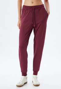Bordeaux joggerbroeken met een elastische tailleband en trekkoord, voorzien van zijzakken en taps toelopende boorden. Matte, gladde stofstructuur.