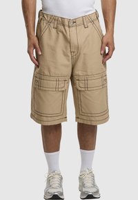 Beige Cargo-Shorts mit sichtbaren Nähten, zwei Vordertaschen und dekorativen Akzenten. Das Material wirkt strapazierfähig und leicht.