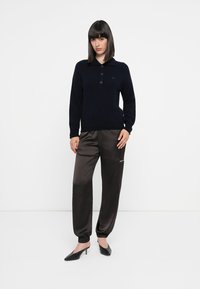 Donna in piedi, che indossa un maglione polo nero a maniche lunghe, pantaloni jogger neri lucidi e mule con tacco neri, su uno sfondo bianco.