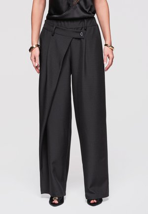 Pantalon noir taille haute à jambes larges avec un panneau frontal asymétrique, détail bouton, porté avec des talons ouverts noirs et des bracelets dorés.