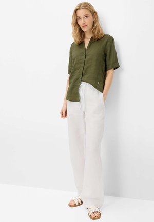 Kvinde står mod en hvid væg iført en olivengrøn kortærmet skjorte, løse hvide bukser og hvide sandaler med spænder.