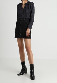 Long-sleeve, navy blouse with a geometric pattern, paired with a black denim skirt featuring buttons. Black patent leather ankle boots.