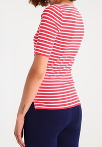 Tričko s krátkým rukávem pruhované v červené a bílé barvě, přiléhavého střihu a zaobleným lemem. V kombinaci s námořnickými modrými kalhotami.