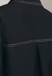 Sort beklædning med kontrasterende hvid syning, der har en struktureret krave og plisseret design bagpå. Glat stoftekstur.
