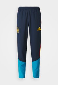 Jūras spēku sporta bikses, kurām ir zils apakšējais segments, dzeltenais Adidas logo, Spānijas nacionālais emblēma un elastīgs jostasvieta komfortam.