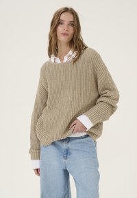 Pulover oversized bej cu rimă, cu un decolteu larg, dintr-un material texturat și cu mâneci lungi, purtat peste o cămașă albă cu guler. Blugi bleu deschis.