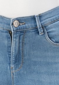 Traperice u svijetloplavoj boji, s patentnim zatvaračem i metalnim dugmetom. Vidljivi su detalji šivanja uz pojas i džepove.