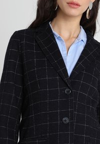 Femme portant des boucles d'oreilles créoles, une chemise bleu clair à col et un blazer noir à motif à carreaux blancs, avec deux boutons attachés.