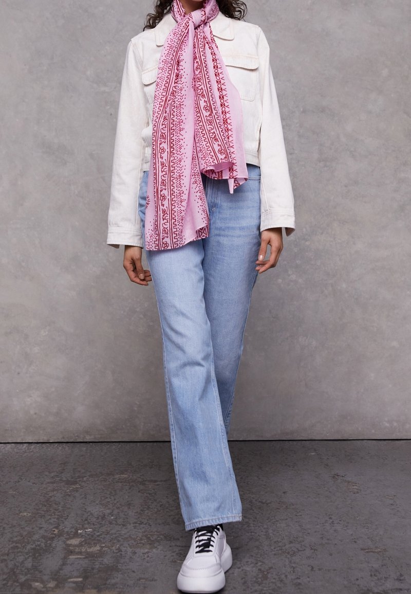 Personne portant une veste blanche, un jean bleu clair, des baskets blanches et une écharpe rose à motifs, debout devant un fond gris texturé.