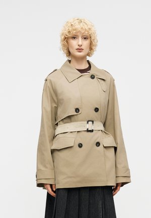 Γυναίκα με κοντά, σγουρά ξανθά μαλλιά, που φοράει ένα μπεζ παλτό τραντσκοτ με ζώνη και μαύρα κουμπιά, συνοδευόμενο από σκούρα πλισέ φούστα, σε απλό φόντο.