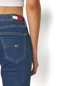 Jeans de mezclilla azul de talle alto con un bolsillo trasero visible, que presenta un pequeño parche bordado del logo en la cintura.