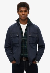 Navy jacket with two chest pockets, zip closure, and folded collar over a green plaid shirt, paired with dark jeans and a watch.
