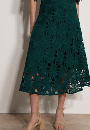 Kobieta w ciemnozielonej, kwiecistej koronkowej spódnicy midi, w połączeniu z beżowymi sandałami na słupku z paskiem przy kostce, stojąca na tle jednolitej ściany.