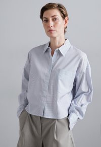 Striped button-up shirt in light blue and white, with a pocket on the left side and long sleeves. Paired with light gray trousers.