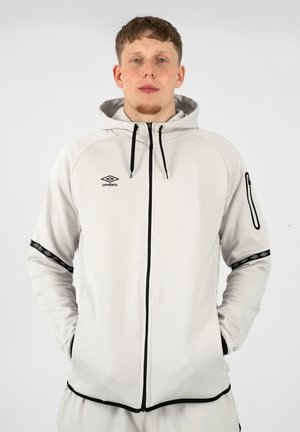 Giovane uomo che indossa una felpa con zip Umbro grigio chiaro con dettagli neri, mani in tasca, in piedi davanti a uno sfondo bianco uniforme.