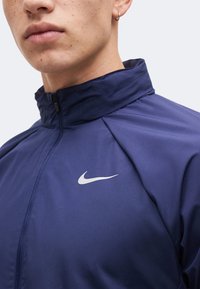 Κοντινό πλάνο ενός ατόμου που φοράει ένα σκούρο μπλε μπουφάν Nike με φερμουάρ και λευκό λογότυπο Nike Swoosh στο στήθος, με ένα μικρό στεφάνη στο αυτί εμφανές.