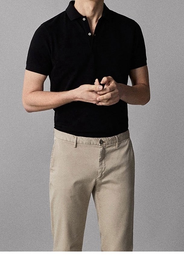 Personne portant un polo noir à manches courtes et un pantalon beige, les mains jointes devant un fond gris uni.