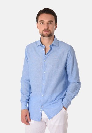 Uomo con barba indossa una camicia azzurro chiaro con bottoni e pantaloni bianchi, in piedi con una mano in tasca davanti a uno sfondo uniforme.