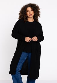 Langer schwarzer Cardigan aus weichem Stoff, offenes Frontdesign, lockere Passform, mit zwei seitlichen Taschen, getragen über einem schwarzen Top und blauen Jeans.
