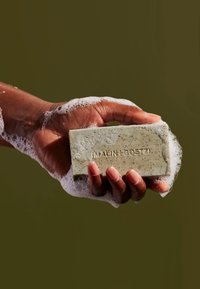 Eine Hand hält einen rechteckigen Block grüner Peeling-Seife mit schwarzen Sprenkeln, teilweise mit Blasen bedeckt, auf dem der Markenname eingeprägt ist.