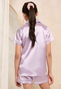 Set di pigiama in raso lilla con maniche corte e pantaloni corti, caratterizzato da dettagli in piping bianco. Il tessuto ha una texture liscia e lucida.
