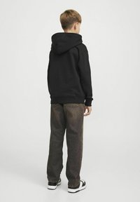 Sudadera de algodón negra con capucha ajustable, combinada con pantalones de mezclilla marrón deslavado y zapatillas negras y blancas, vista desde la parte de atrás.