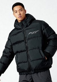 Zwarte donsjas met een hoge kraag, ritssluiting en de tekst 'volg je hart'. Dikke doorgestikte stof, geribbelde manchetten en zoom.