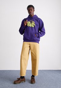 Paarse hoodie met grafische print, beige wijde broek en bruine veterschoenen. Casual kleding met een textuurafwerking en een losse pasvorm.