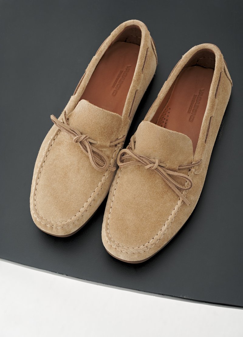 Een paar beige suède loafers met decoratieve strikveter, rustend op een donkere, gladde ondergrond.
