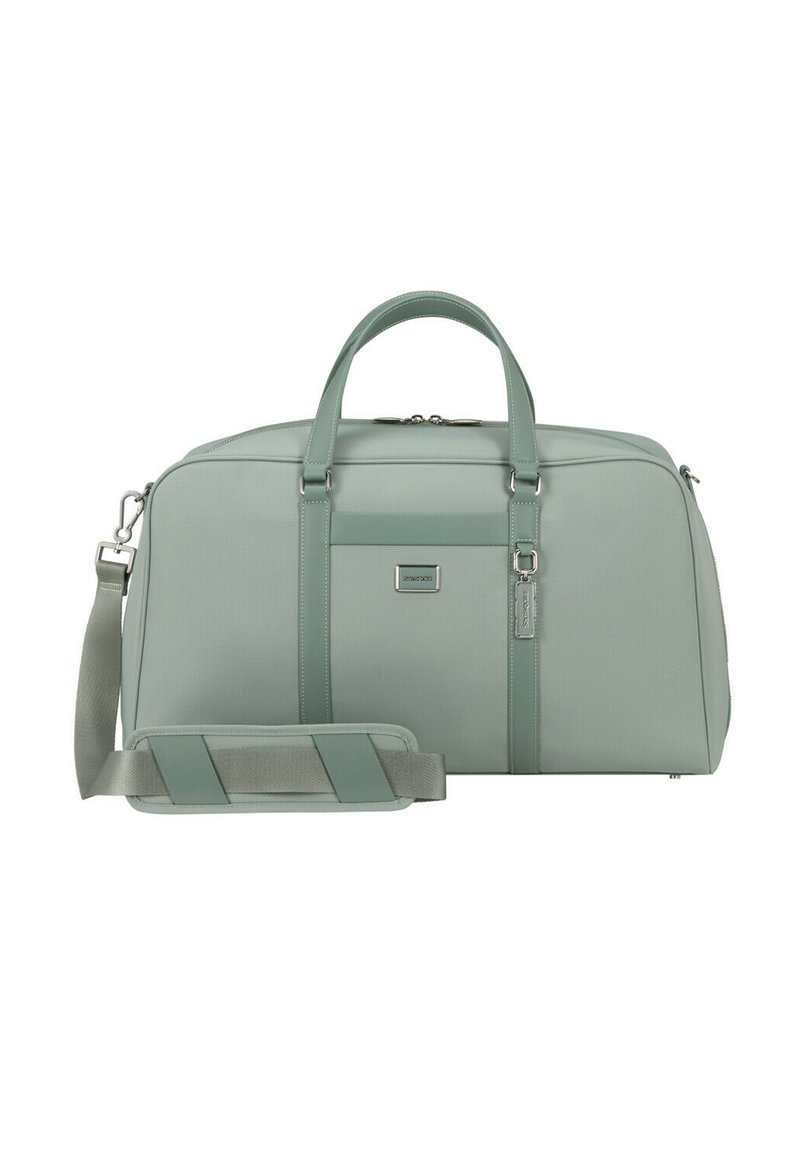 Erweiterbare Reisetasche aus weichem mintgrünem Stoff, mit zwei oberen Griffen, einem abnehmbaren Schultergurt und einem Logo-Patch auf der Vorderseite. Silbernene Akzente.