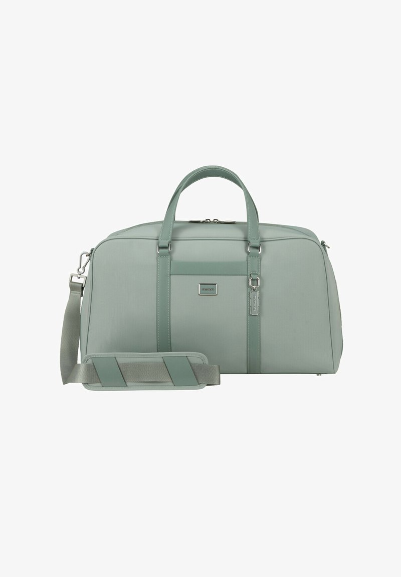 Erweiterbare Reisetasche aus weichem mintgrünem Stoff, mit zwei oberen Griffen, einem abnehmbaren Schultergurt und einem Logo-Patch auf der Vorderseite. Silbernene Akzente.