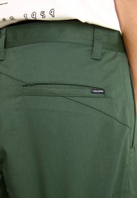 Pantaloni verzi închis cu o textură netedă, având un buzunar pe spate și un mic eticheta cu logo-ul Volcom. Croială modernă cu linii curate.